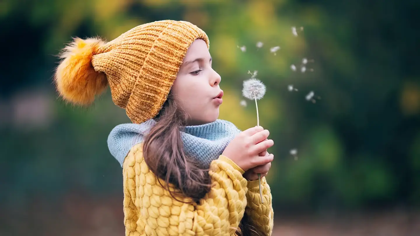 A young girl standing outside blowing a dandelion