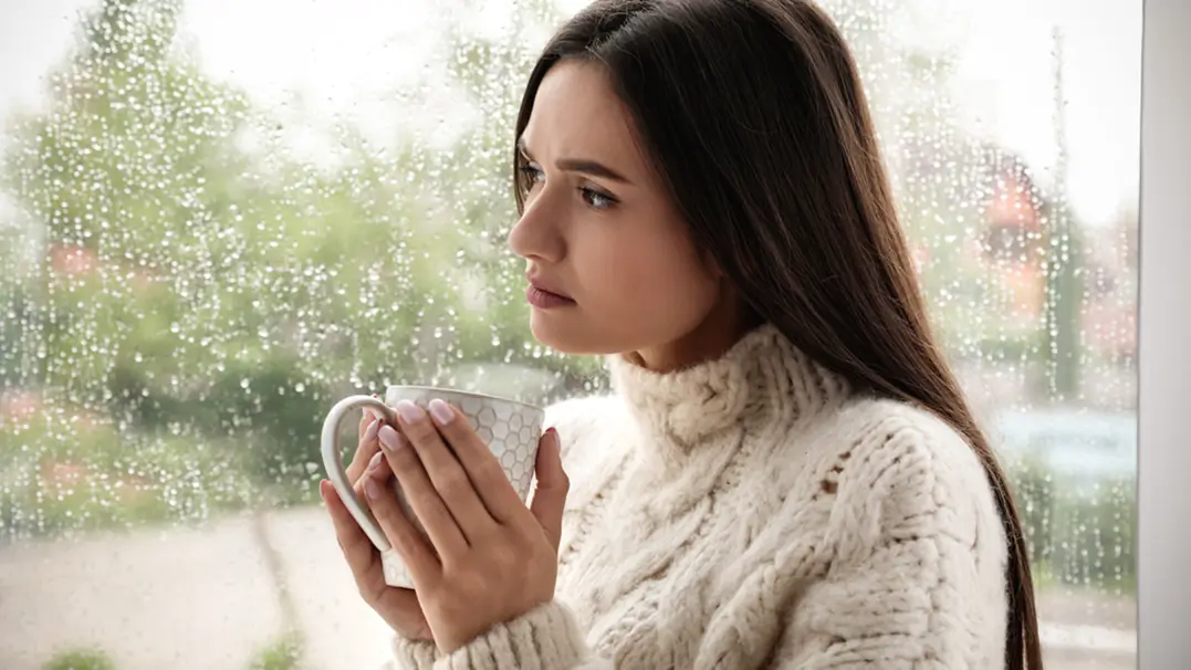 A woman standing next to a window enjoying a drink out of a mug.